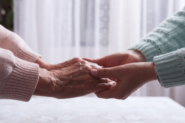 An older woman’s hands are held by a younger woman’s hands, symbolizing mental health concerns in an aging parent.