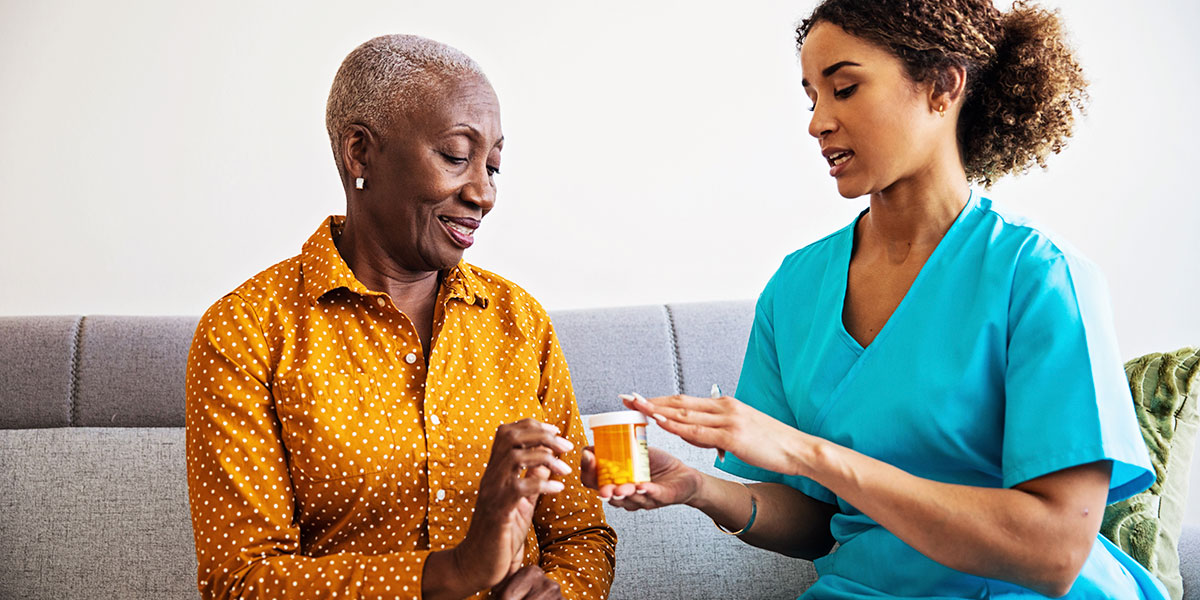 A nurse providing medication management services holds a prescription pill container and a clipboard as she talks with an older woman.