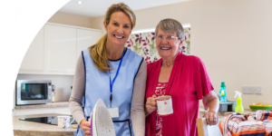 A caregiver providing senior housekeeping services in Saginaw holds an iron and smiles beside the older woman she is helping.