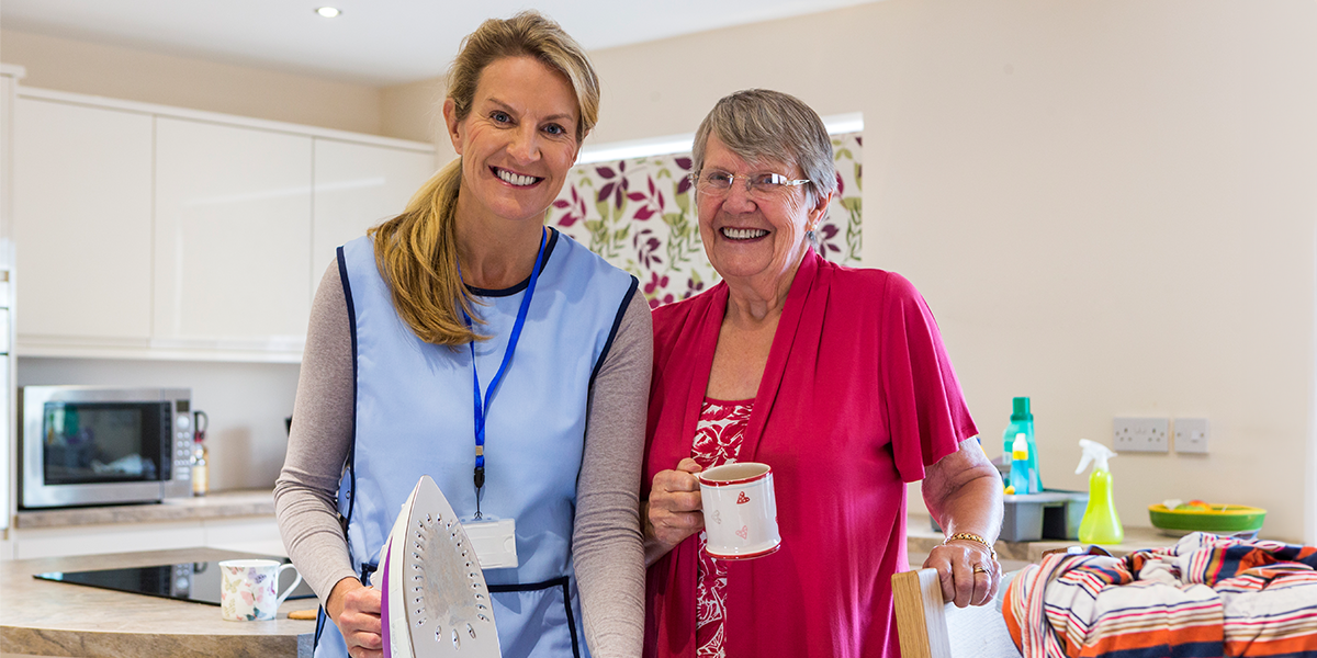 A caregiver providing senior housekeeping services in Saginaw holds an iron and smiles beside the older woman she is helping.