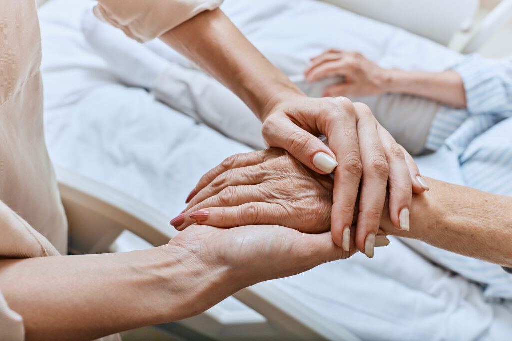 A younger woman’s hands hold the hand of an older woman as she lies in bed, displaying signs of the last stage of dementia.