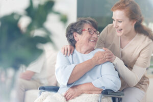 : A caregiver providing a home care solution for aging adults gives an older woman in a wheelchair a hug and a smile.