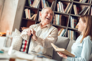 An older man who has learned how to maintain independence with MS shares a laugh with his caregiver as she reads to him from a book.